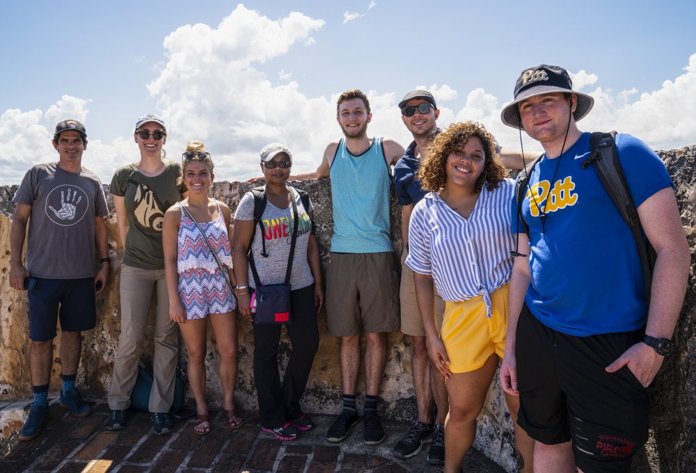 Students pose for a group picture in front of a wall in old San Juan with bright sunny skies and clouds overhead